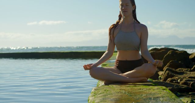 Young Caucasian woman practices yoga by the sea. Her serene pose on the rocky shore suggests a peaceful meditation session outdoors.