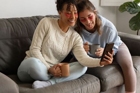 Smiling diverse women relaxing with under-eye hydrogel patches and coffee while sharing phone