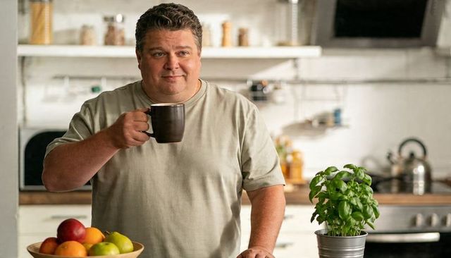 Man holding coffee mug at kitchen counter with fresh basil and fruit bowl, casual morning