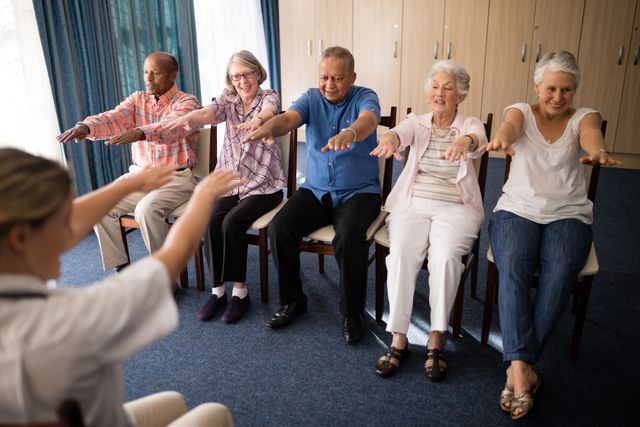 Senior group participating in a fitness session with a female doctor in a retirement home. Ideal for use in healthcare, senior care, wellness programs, and physical therapy promotions. Highlights active seniors and the importance of maintaining health and fitness in older age.