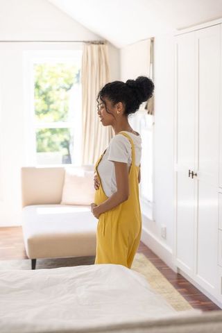 Pregnant woman in yellow overalls standing in sunlit airy bedroom, serene maternity moment