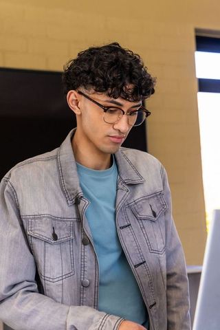 Young professional concentrating on laptop wearing denim jacket and round eyeglasses