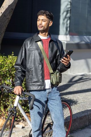 Asian man standing with bicycle holding mobile wearing leather jacket and green sling bag