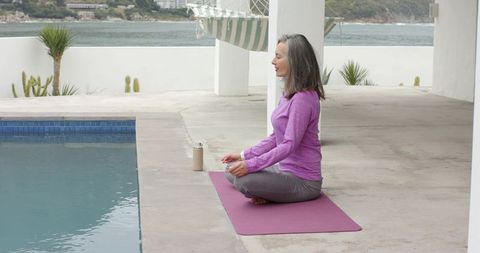 Senior woman meditating cross-legged on purple yoga mat by pool with coastal view