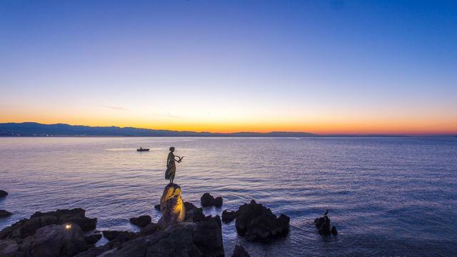 Statue of a woman stands on a rock at the seashore during sunset. Calm sea with a small boat in the distance and dramatic twilight colors on the horizon creates a tranquil atmosphere. Perfect for travel brochures, serene landscapes, or nature themed projects.