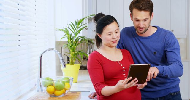 Caucasian man and Asian woman review a tablet in the kitchen. They're collaborating on a recipe or planning with a digital device at home.