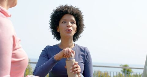 African american woman opening reusable bottle while chatting on coastal terrace