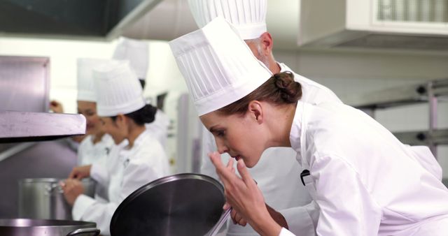 Chefs dressed in traditional white uniforms and hats are working together in a commercial kitchen. One chef is lifting the lid of a pot and inspecting the contents, while others are focused on their cooking tasks. This image can be used for articles related to culinary schools, professional cooking, team collaboration in kitchens, and the culinary profession.