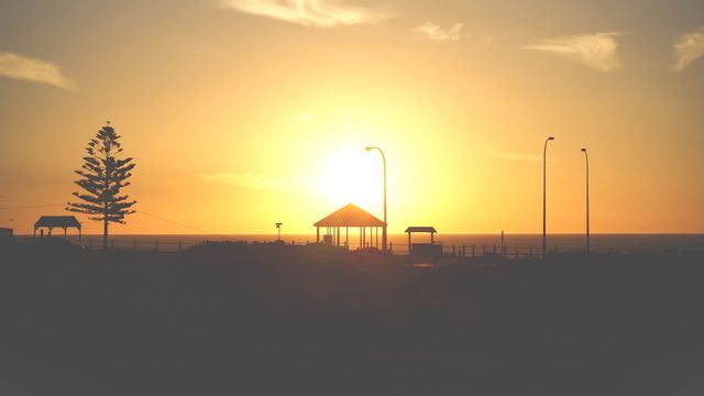 Golden hour sunset casting silhouettes of a gazebo and trees at a serene coastal beach. Perfect for travel magazines, nature blogs, and landscape calendars to evoke relaxation and the beauty of seaside sunsets.