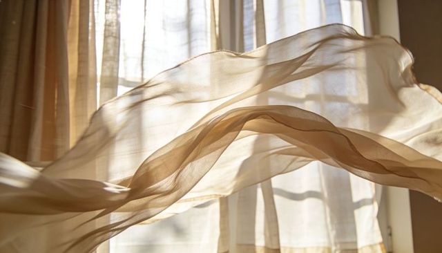 Sunlit sheer beige curtains billowing over window with layered voile drapery