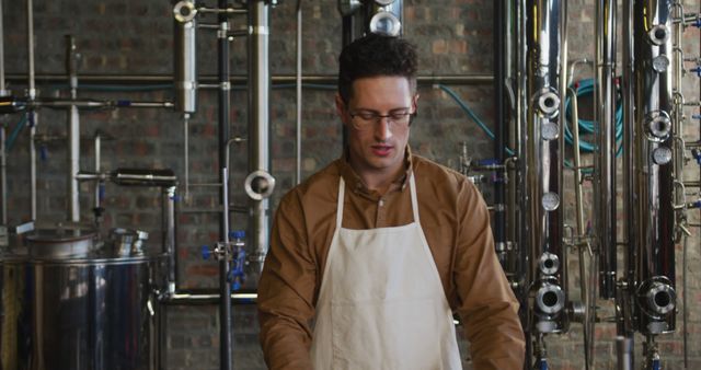 Man in an apron, wearing glasses, working in a craft brewery facility. He is surrounded by industrial brewing machinery. This image is ideal for use in articles or advertisements related to craft beer, brewing processes, beverage manufacturing, or industrial production. It can also be used for topics on small business operations or profiles on industry professionals.