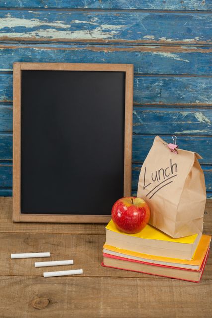 School supplies including a chalkboard, chalk, an apple, a lunch bag, and books are arranged on a wooden table against a blue wooden background. This image is ideal for educational materials, back-to-school promotions, and classroom decor.