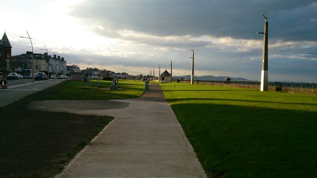 Empty coastal pathway lined with grass and streetlights, extending towards horizon, at dusk with cloudy sky. Ideal for themes of tranquility, evening walks, urban coastlines, sunset ambiance, peaceful environment, and residential seaside life.
