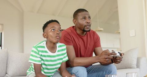 African american father and son playing video games on living room sofa, bonding