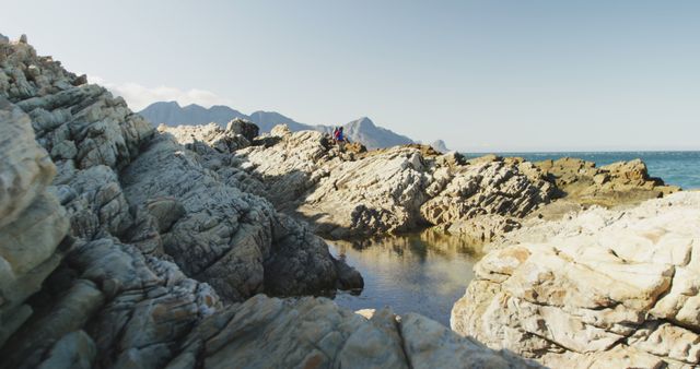 This image captures a breathtaking rocky coastal landscape with mountains in the background and a calm sea under a clear sky. Perfect for travel magazines, nature blogs, outdoor adventure promotions, and background for websites focusing on natural beauty or tranquility.