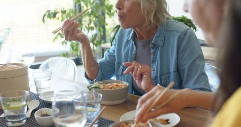 Mature friends enjoying casual asian-inspired lunch at sunlit dining table using chopsticks