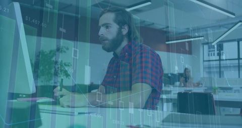 Bearded man analyzing financial charts on desktop while writing on tablet in open office