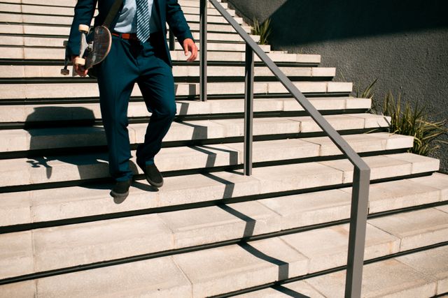 Businessman in a suit holding a skateboard while walking downstairs. This image can be used to depict modern business lifestyles, active professionals, or creative work environments. Ideal for articles or advertisements focusing on work-life balance, urban mobility, or innovative business practices.