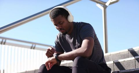African American Runner Checking Smartwatch Resting on City Steps Wearing Headphones