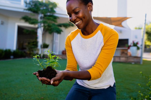 African American woman holding a plant seedling with soil in her hands while smiling in a garden. Ideal for use in content related to gardening, sustainability, environmental awareness, outdoor activities, and lifestyle blogs. Perfect for illustrating concepts of growth, nature, and personal hobbies.