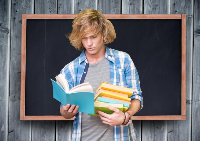 Teenage student reading a book while holding several others, standing in front of blackboard. Use for educational content, school promotions, learning resources, academic advertisements, or study tips.