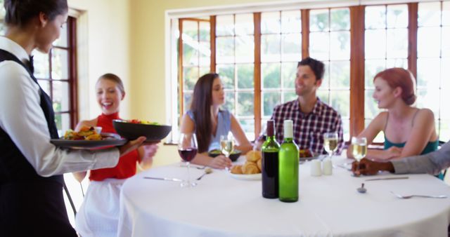 A group of friends is sitting around a table enjoying a meal in a well-lit restaurant. The waiter is serving food, and there are glasses of wine on the table along with various dishes. The setting suggests a social gathering or celebration. This image is ideal for use in advertising for restaurants, social events, dining experiences, and hospitality services.