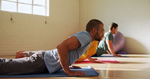 Three people in a sunlit studio practicing yoga on mats while performing the cobra pose. Ideal for use in concepts of group exercise, yoga classes, healthy lifestyle, fitness routines, wellness promotions, and indoor physical activity marketing materials.