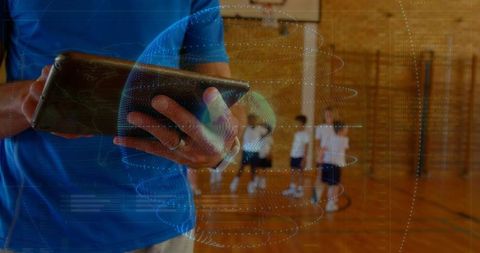 Coach holding tablet with holographic globe overlay supervising children in school gym