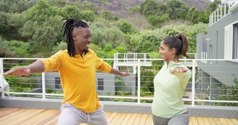 Multicultural couple practicing warrior ii yoga on wooden deck with greenery and modern home