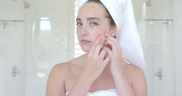 Young woman focusing on facial skincare routine in bathroom. She is treating acne by examining her skin in the mirror. Ideal for content related to skincare, acne treatment, beauty routines, personal care products, and hygiene tutorials.