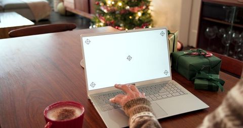 Mature woman typing on laptop amid holiday gifts and cozy cocoa by christmas tree