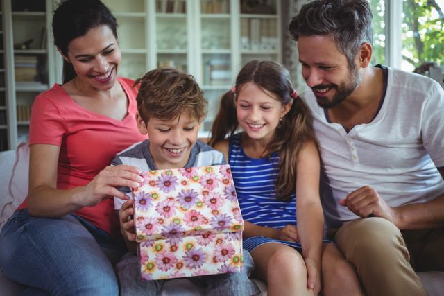 Family sitting on couch, opening a gift box with floral wrapping paper. Parents and children smiling and enjoying the moment. Perfect for themes related to family bonding, celebrations, parenting, and joyful moments at home.