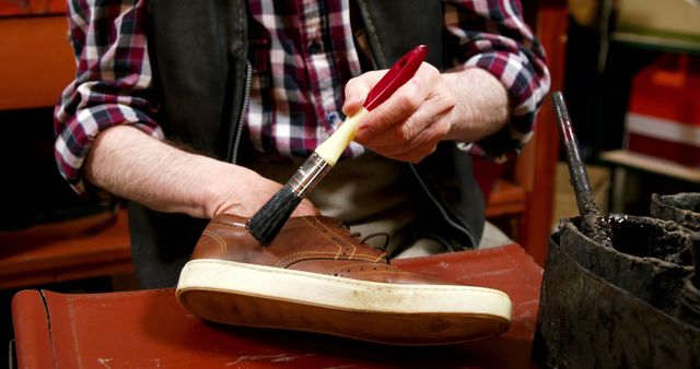 This vibrant stock photo of a cobbler polishing a brown casual shoe can be used to represent traditional craftsmanship, shoe maintenance, and skilled labor. Ideal for blogs, articles, and commercials focusing on shoe care, vintage trades, and hardworking artisans. It highlights the use of simple tools, like a paintbrush, and can be perfect for illustrating the detailing work done in shoe repair shops.