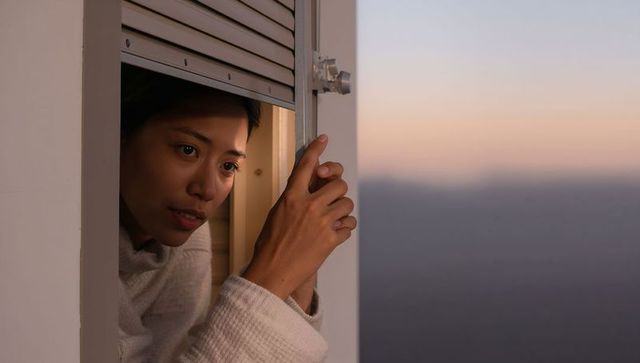 Contemplative asian woman leaning at window shutter, peering into twilight with warm glow