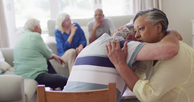 Elderly friends are consoling each other in a living room during a group discussion. Other friends are talking in the background. This can be used for topics related to senior care, emotional support, friendship, and community among elderly people.