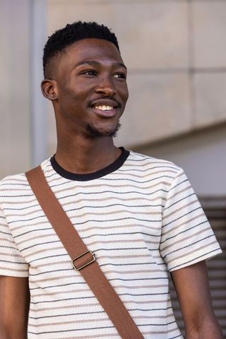 Smiling african man wearing striped shirt and brown crossbody bag at urban facade