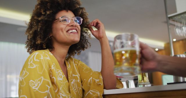 Woman with curly hair and glasses smiling while sitting at a pub. She is wearing a yellow shirt and enjoying a beer offered by someone else. This image can be used to depict socializing, relaxation, happiness, pub culture, and leisure activities.