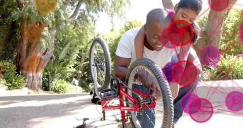 Dad repairing upturned red bicycle on sunny driveway while daughter watches and smiling