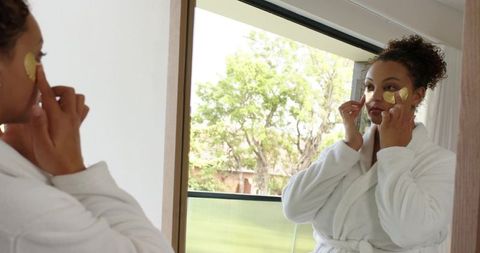 African american woman applying gold under-eye patches wearing white robe by balcony window