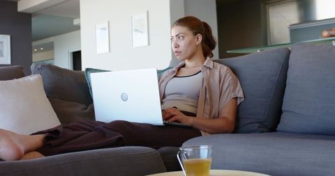 Young woman working on laptop at home on cozy grey sofa with blanket and tea cup minimal decor