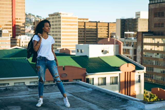 Front view of a hip young biracial woman wearing jeans and white trainers, holding a leather jacket standing on an urban rooftop with buildings in the background.