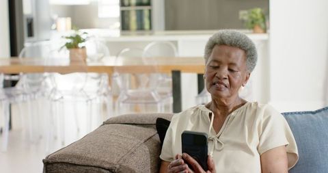 Senior african american woman using smartphone on bright modern living room sofa, relaxed