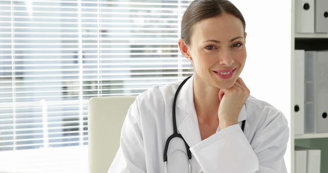 This image of a smiling female doctor in a white coat with a stethoscope in her office is perfect for medical and healthcare advertisements, clinic websites, patient education materials, and articles on healthcare professionals. The professional setting and confident expression convey trust and competence in medical services.