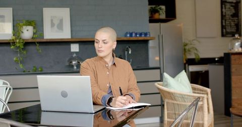 Shaved-head professional working on laptop and taking notes in modern cozy kitchen cafe