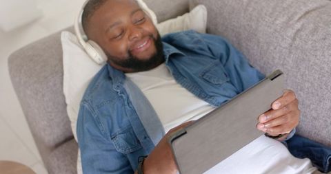 African american man reclining on couch relaxing with tablet and wireless headphones