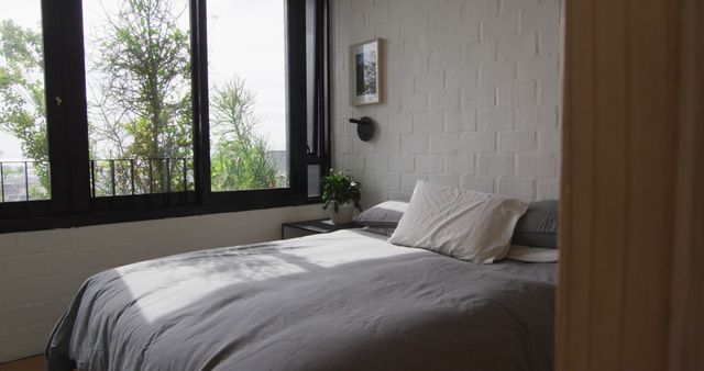 Cozy bedroom features brick walls and large windows that allow ample natural light to fill the space. A comfortable bed is adorned with simple bedding, complemented by an indoor plant and minimalist decor. Ideal for depicting modern urban living, relaxation, and home comfort themes in digital and print media.