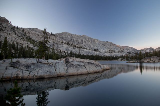 Image captures quiet mountain lake at dawn, showcasing calm, reflective waters surrounded by rocky terrain and sparse trees. Ideal for use in nature travel brochures, meditation and relaxation materials, environmental campaigns, and scenic calendars. Offers a feeling of peaceful solitude and untamed beauty.