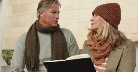 Mature couple sitting on bench wearing cozy knit scarves and beanie holding open notebook