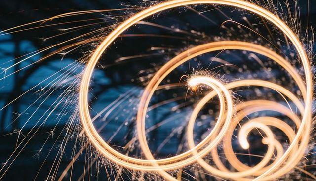 Golden sparkler spiral trails creating glowing rings and flying sparks against night bokeh