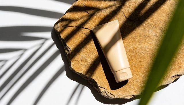 Beige cosmetic tube resting on textured stone slab casting palm-leaf shadows